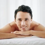 A man lays on his front on a treatment bed at a beauty salon, he rests his chin on his hands and smiles